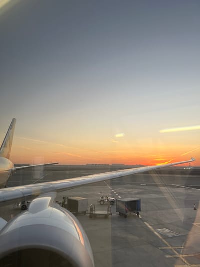 A rotated view from an airplane window depicts the wing and engine against a sunset sky. The terminal tarmac with baggage carts is visible beneath the wing. The sky transitions from dark on the left to shades of orange and yellow near the horizon. The plane's white and gray wing and engine dominate the lower right, while the wing extends to the top of the frame. A bright reflection distorts the view.
