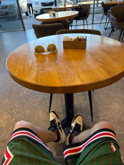 A person wearing green and red basketball shorts sits at a round wooden table with a black metal frame. The table is located in an outdoor area of a cafe or restaurant. The table has a small brown box with sugar packets on it, and white sunglasses are resting on the table's surface. The floor is made of gray tiles, and there is a glass door to the left of the table. There are other tables and chairs in the background, but they are out of focus. The person's feet are resting on the floor, and they are wearing white and black sneakers. Based on the provided coordinates, the location of the image is in the city of Kokhav Ya'ir Tzur Yigal, Israel.