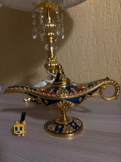 The scene is set indoors in Mikrevo, Bulgaria. A highly ornate, decorative genie lamp, finished in gold with extensive blue and red enamel detailing, rests on a light wood-grain surface in the foreground. To the immediate left of the lamp, a small, blocky yellow figurine with black markings and green eyes, consistent with a Minecraft-style ocelot or cat, features a black attachment loop on its back. In the blurred background, an illuminated golden table lamp, characterized by a white lampshade and multiple clear crystal drop embellishments suspended from its central column, is situated against a plain, light beige wall. No people are visible.