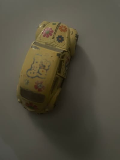 The image shows a yellow toy car with floral designs and the word "Go Go" on it. The car is positioned on a flat, light-gray surface, likely indoors, and appears to be at an angle relative to the viewer. A soft shadow is visible on the right side of the image. The location in the city of Ebo, Nigeria is unknown.