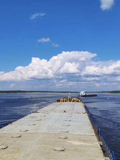 The image depicts a wide, flat barge traveling on a wide river under a partly cloudy blue sky. The barge deck is grey and has numerous circular access panels. Blue railings are visible along the edges of the barge. In the distance, another vessel, a ferry barge, is visible further up the river. Tree-lined riverbanks are seen in the background and there are electrical transmission towers in the far distance. The location is likely near Novodevich'e, Russia, based on the presence of large river systems, similar infrastructure, and general landscape features. No people are visible.