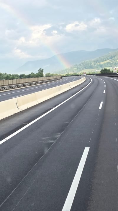 A vertical, eye-level shot captures a highway curving to the right under a cloudy sky where a faint rainbow is visible. The highway is paved and has white lane markings. A concrete barrier separates the lanes from a raised median with a metal guardrail. Green trees and hills form the background, with a faint rainbow arcing across the sky above them. The scene appears to be located on a highway near Au bei Gaishorn am See, Austria.
