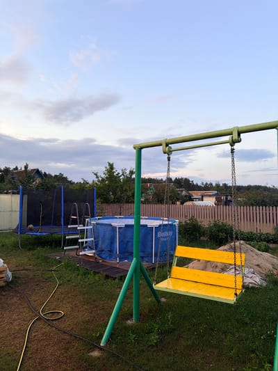 A wide shot captures a backyard scene in Радужскі сельскі Савет, Belarus, at dusk. A yellow and green swing set stands in the foreground, with a yellow wooden seat hanging from metal chains. To the left, a blue inflatable swimming pool with a ladder is partially visible, situated next to a trampoline. Beyond these play structures, a wooden fence separates the yard from a residential area with houses and trees under a pale blue sky with scattered clouds. Yellow and black hoses lie coiled on the grass.