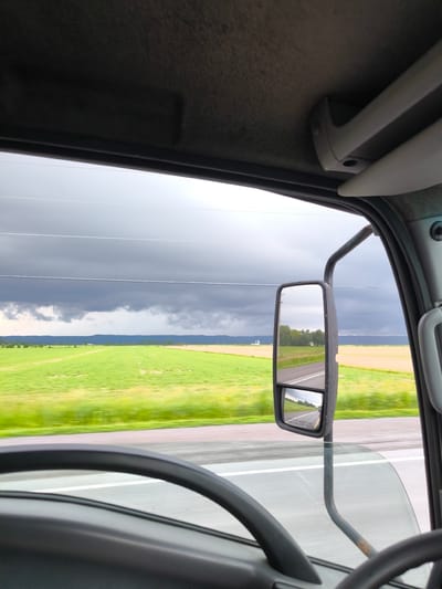 The image provides an interior view from the cab of a vehicle, looking out at a rural landscape. The foreground is dominated by the dark interior of the vehicle, including a portion of the steering wheel and the dashboard. A large side mirror is prominently featured, reflecting the scene behind the vehicle.
Outside the vehicle, the scenery consists of vast green fields stretching towards a distant horizon. The sky above is overcast with heavy, dark clouds, suggesting impending or ongoing rain. The weather appears to be gloomy and possibly stormy. The fields are a vibrant green, indicating a lush agricultural setting. In the distance, there are faint outlines of trees and perhaps some small buildings or structures.
The side mirror shows a reflection of the road and the surrounding landscape. This reflection also captures the dark, cloudy sky. The overall mood of the image is somewhat somber due to the weather, but also evokes a sense of travel through open country. The location is identified as Champlain, Canada, which aligns with the rural agricultural scenery depicted. No people are visible in the image. There is no discernable text in the image.