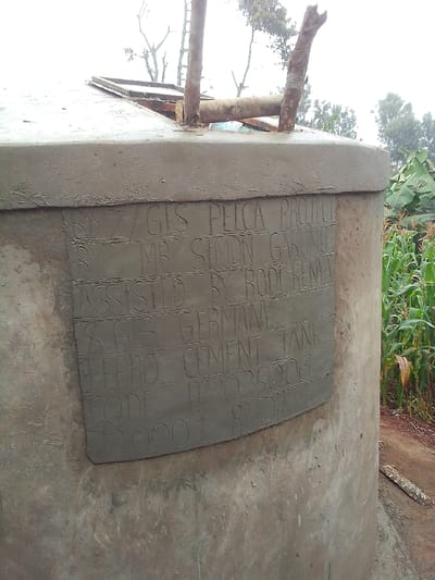 The image displays a concrete cylindrical water storage tank situated in Ihururu, Kenya. The tank features a flat concrete top with a rectangular metal access hatch located towards the left side. Two vertically oriented, debarked wooden poles are embedded into the top surface, along with a horizontal wooden branch resting upon it. On the tank's front-facing side, a rectangular panel of lighter gray cement is visible, containing incised text in multiple lines. The legible text states: "BNZ / GIZ PELCA PROJECT," "BY MR. SIMON GARUNYU," "ASSISTED BY RODA KENYA," "GIZ GERMANY," "FERRO CEMENT TANK," "CODE FC2025006," and "12000 L BY [partially obscured]". The main structure of the tank is composed of light gray concrete. To the immediate right of the tank, a dense growth of tall, green maize (corn) plants is present. The ground visible around the tank is reddish-brown soil. The background consists of blurred green foliage and trees, set against an overcast or bright sky. No individuals are present in the scene.