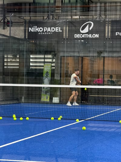 In Ban Buak Khrok North, Thailand, a young man in athletic attire is captured mid-swing on a vibrant blue padel court. He wears a light-colored sleeveless shirt, dark shorts, and white sneakers with black accents. The court is dotted with several yellow padel balls, some scattered near the net. A white net divides the court. Behind the net, the backdrop features glass walls with various advertisements and branding, including "Niko PADEL ACADEMY" and "DECATHLON". Indoor plants in white pots are visible, adding a touch of greenery to the setting. Reflections on the glass indicate an indoor environment.