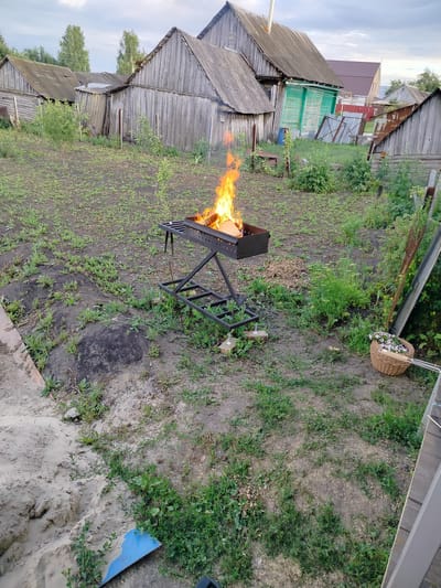 The image depicts a rural outdoor scene in the village of Vazery, Russia. In the foreground, a metal brazier is alight with a fire, with pieces of wood visible within. The brazier is situated on a grassy, uneven area with patches of bare earth. To the right of the brazier, a woven basket filled with white flowers sits on the ground, next to a metal pole. Further into the background, several weathered wooden structures, likely sheds or outbuildings, are visible. A house with a red roof and green siding is also present. The sky is overcast with a hint of clouds. The ground is covered with grass and dirt, with some sand visible in the lower left corner.