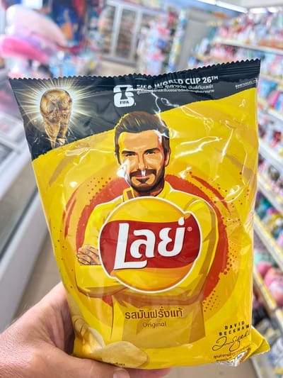 This is a close-up, slightly angled shot of a person holding a bag of Lay's potato chips. The bag is predominantly yellow and features an illustration of David Beckham, with the FIFA World Cup 2026 branding prominently displayed. The text on the bag is in Thai and English, indicating the flavor is "Original" and celebrating the upcoming World Cup. The scene appears to be inside a store, with shelves of other products visible in the blurred background. The lighting is bright and suggests an indoor environment. The person's hand holding the bag is the only visible part of them. The focus is entirely on the product and its promotional design. The background suggests a retail setting, possibly a convenience store or supermarket aisle. The overall impression is a commercial advertisement for a limited edition product.