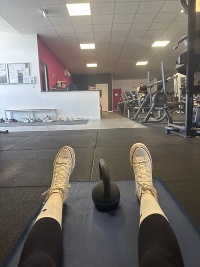 The image depicts a first-person view from a gym in Neuville-aux-Bois, France. In the foreground, two legs are extended forward, clad in black athletic pants, white socks, and white Converse high-top sneakers with red trim on the sole. The feet rest on a dark blue yoga mat, which is positioned on a black rubber gym floor composed of interlocking square tiles. Between the feet, a black kettlebell with a blue base rests on the yoga mat.
The midground features an open expanse of the black rubber gym floor. To the left, a light-colored bench holds multiple pairs of casual shoes. Beyond the bench is a white reception desk with a vibrant pink wall behind it. An air conditioning unit is mounted on the pink wall, and a bulletin board with various papers is visible on the adjacent white wall.
The background, primarily to the right of the frame, shows a diverse array of gym equipment, including weight machines, racks, and barbells, extending into the distance. One individual is visible operating a piece of cardio equipment in the far background. The entire space is illuminated by bright, square fluorescent lights set into a white grid ceiling. The walls in the background feature sections of pink and dark grey paint.