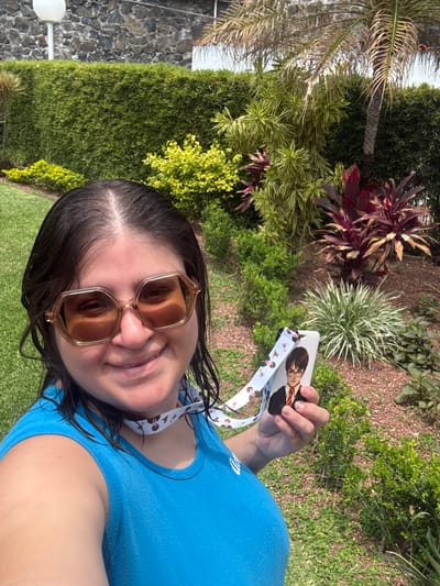 A woman is taking a selfie outdoors, holding up a lanyard and ID card with an image of Harry Potter. She is wearing sunglasses and a blue t-shirt. The background consists of lush green foliage, including bushes, grass, and various plants. A stone wall and a lamppost are also visible in the background. The location appears to be in a well-maintained garden or park. The city is Tres de Mayo, Mexico.