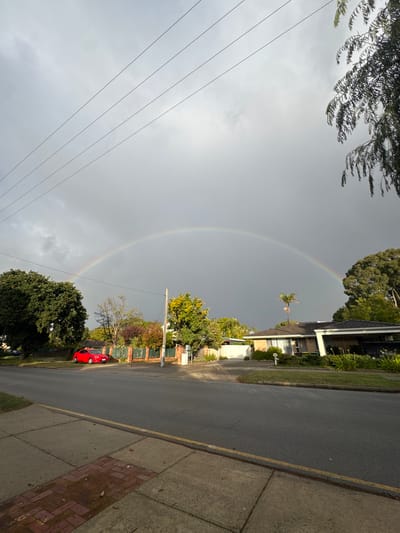 The image depicts a residential street scene with a rainbow in the sky. The location is Bassendean, Australia. A paved road runs along the right side of the frame, with a sidewalk on the far right. Houses are visible on the right side of the street, surrounded by trees. A red car is parked on the side of the road. Overhead power lines stretch across the left side of the frame, partially obscuring the sky. A rainbow arcs across the sky, appearing to end near the houses on the right. The overall weather appears overcast.