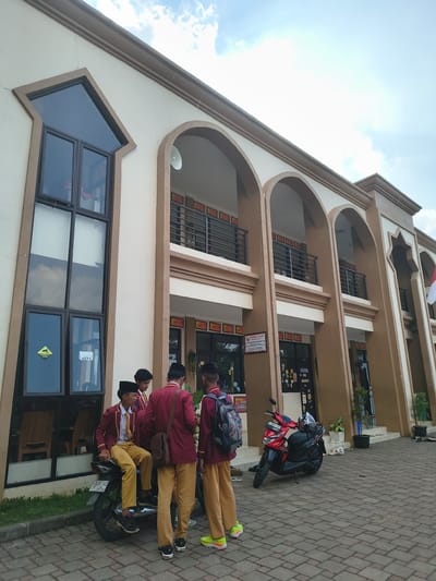 Four young males, dressed in school uniforms consisting of maroon blazers, white shirts, yellow ties, and khaki pants, are gathered outside a building in Ciputri, Indonesia. One individual sits on a black motorcycle. Another, wearing a black peci cap, stands beside him. The third and fourth individuals stand behind them, one with a backpack. The building features a cream-colored facade with brown archways and balconies. A red motorcycle is parked on the paved ground. The sky above is partly cloudy.