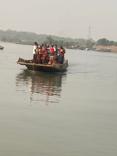 A small boat, heavily laden with numerous passengers, navigates a wide river. The water reflects the boat's hull and the figures aboard. Hazy vegetation lines the riverbank on one side, while a transmission tower is visible in the distance. The boat appears to be the primary mode of transport across the water, emphasizing its importance to the people.