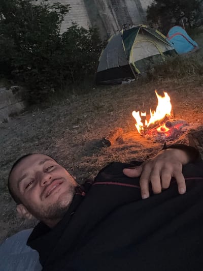 A person is lying down outdoors near a campfire and a tent. The image is taken from a low angle, with the person's head near the bottom of the frame, looking up at the camera. They are wearing a dark jacket. The campfire is burning brightly, casting a warm glow on the surrounding dry grass and dirt. Two tents are visible in the background, one green and grey, and another blue and grey. The scene appears to be set in a rural or camping area, likely during the evening or night due to the campfire and dim lighting. The person's expression is relaxed and friendly. There are no visible text elements in the image.