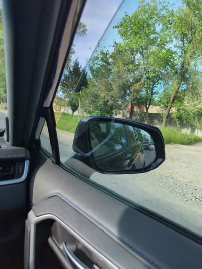 The image shows the interior of a car, focusing on the driver's side window and side mirror. The view through the window includes trees, a blue sky with clouds, and a glimpse of a gravel road. The side mirror reflects the same road and trees, with a vehicle following behind. Inside the car, the door panel is visible, featuring gray upholstery with blue stitching and a silver door handle. A portion of the car's dashboard and controls is also visible. The location is likely Beş-Küŋgöy, Kyrgyzstan, based on the context.