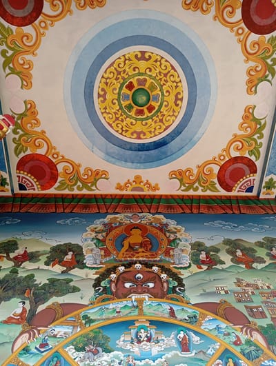 A painting on a ceiling in Lumbini Sanskritik, Nepal, showcases Buddhist imagery. At the top is a large central circle with blue concentric rings surrounding a yellow and green mandala. Decorative scrollwork patterns in yellow and green fill the corners above a band of green, red, and tan geometric shapes. Below this is a blue landscape with small figures seated under trees and a town in the distance. A deity with a red face and three eyes, adorned with skulls, is centrally positioned beneath a seated Buddha figure. Below the deity is a segmented wheel with scenes depicting various figures and actions.