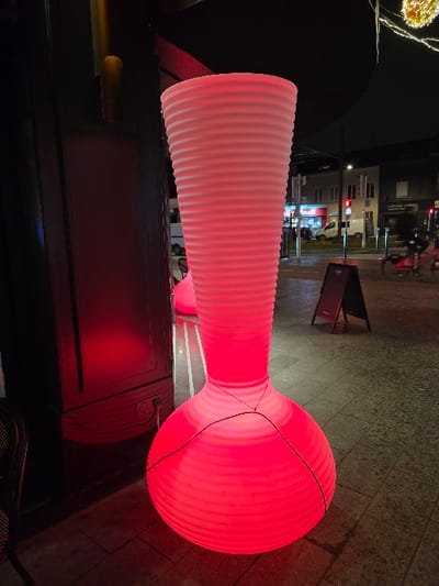 A close-up shot shows a red, hourglass-shaped light fixture. It features a narrow, ribbed cylindrical section that expands to a large, round sphere. The light is positioned near a window, casting a red glow on the surrounding surfaces. The outdoor cityscape is visible through the window.