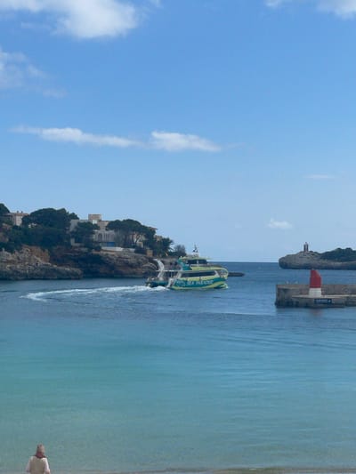 The image depicts a daytime coastal scene in Porto Cristo, Spain, focusing on the mouth of the harbor. The sky is clear blue with scattered white cumulus clouds.
In the mid-ground, a green and yellow passenger catamaran named "SEA PARADISE" with "CLASS BOTTOM CAT" visible on its side, is navigating right-to-left, leaving a white wake in the turquoise water. The boat is positioned between the left-side cliffs and the right-side breakwater.
To the right, a concrete breakwater extends into the water, featuring a prominent red conical navigation beacon or small lighthouse at its end. A sign on the breakwater reads "CLUB NÀUTIC PORTO CRISTO". Two white birds are perched on the breakwater near the red beacon. Beyond the breakwater, a rocky islet with a small white lighthouse structure is visible in the distance, marking the entrance to the open sea.
The left side of the scene is dominated by rocky cliffs covered with green pine trees. Several white-painted buildings and houses are situated atop these cliffs, overlooking the water, with one larger building featuring prominent arches.
In the foreground, a sandy beach with shallow, clear water occupies the bottom of the frame. A single individual, viewed from behind, stands at the water's edge or in very shallow water. The person is wearing a light-colored, possibly beige, top, a dark reddish scarf around their neck, and a light-colored knitted cap. No specific actions or interactions are discernible for this individual. The water gradually deepens and changes to a darker blue hue towards the horizon.