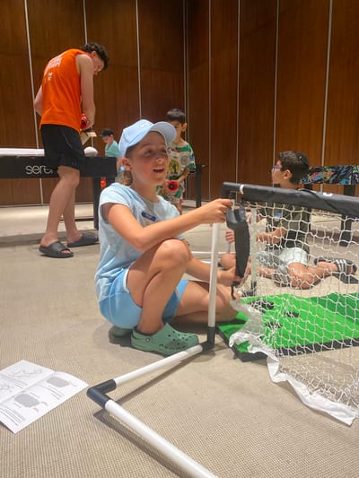 A wide-angle, medium shot taken indoors shows a group of children engaged in activities. A boy wearing a light blue shirt, matching shorts, a light blue baseball cap, and green Crocs is seated near a small, portable soccer goal with a white and green net. He is holding onto the goal's frame. To the left, another child in an orange shirt and black shorts is holding onto a black structure with white lettering. Other children are visible in the background. A set of assembly instructions lies on the carpeted floor near the goal. The environment suggests an indoor recreational setting. The location, Marina Maroma, Mexico, is not discernible from the visual information alone.