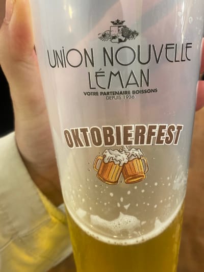 A close-up, eye-level photograph depicts a transparent plastic cup containing approximately 0.5 liters of a golden-yellow, carbonated beverage identified as beer, topped with a substantial layer of white foam. The cup is held by a person's left hand, with the thumb and index finger visible on the left side. The individual is wearing a light-colored, possibly beige or yellow, long-sleeved garment.
The cup's exterior features prominent branding. At the top, a crest-like emblem is present, followed by the text "UNION NOUVELLE LÉMAN" in large capital letters. Below this, "VOTRE PARTENAIRE BOISSONS DEPUIS 1936" is inscribed. Further down, the word "OKTOBIERFEST" is displayed in bold, brown block letters, beneath which is an illustration of two traditional, clinking beer steins with overflowing foam. Small bubbles are visible clinging to the interior surface of the cup.
Union Nouvelle Léman is a known beverage distributor based in Thonon-les-Bains, France. The presence of the "OKTOBIERFEST" branding indicates the cup is from an Oktoberfest-themed event, likely held within Thonon-les-Bains or its immediate vicinity. The background is out of focus, suggesting an indoor setting, possibly an event venue or establishment.