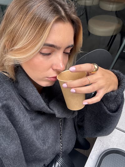 A young adult female is depicted in a close-up, high-angle shot, bringing a plain, light brown paper cup to her lips. Her eyes are closed, and her head is tilted slightly downward, indicating focus on the object or its contents. She has blonde hair with darker roots, parted on the side, and well-defined dark eyebrows. Her skin is light-toned, with visible cosmetic enhancement around the cheeks. She is wearing a dark gray, textured, cowl-neck garment, possibly a sweater or coat. Her left hand, adorned with light pink nail polish, holds the cup. A large, polished gold-colored domed ring is on her left ring finger, and a thin, gold-colored band is on her left middle finger. A black quilted bag with a silver-colored chain strap is partially visible below her left arm. The background includes reflections of circular light-colored objects, possibly stools or tabletops, in a dark, reflective surface, along with a dark chair element. A light gray, textured tabletop and a black rectangular tray or plate are visible in the lower right foreground. The scene is situated in Vaulx-en-Velin, France.