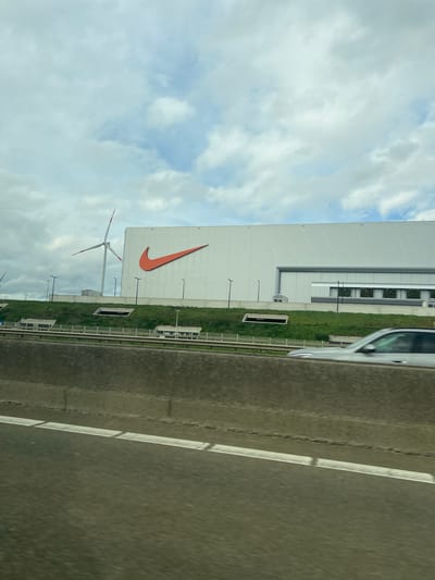 The image shows a white, modern building with a large orange Nike swoosh logo on its side. In front of the building is a field with multiple wind turbines. To the right is a highway.
The image was taken in Kwaadmechelen, Belgium.