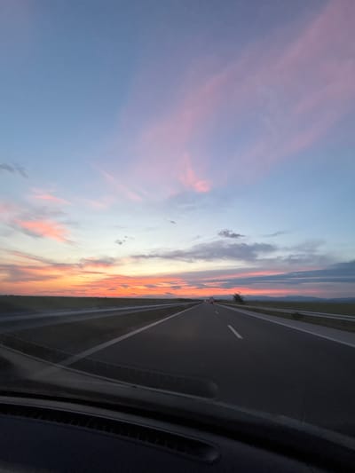 The image depicts a sunset sky with orange and pink hues, visible from inside a car traveling on a highway. The highway has multiple lanes and is marked with white lines. The sky fills the majority of the frame on the left side, while the highway occupies the right side, with the car's interior visible in the bottom right corner. The location is indicated as Градина, Bulgaria, but there are no distinctive features visible in the photograph to confirm the exact spot within the city.