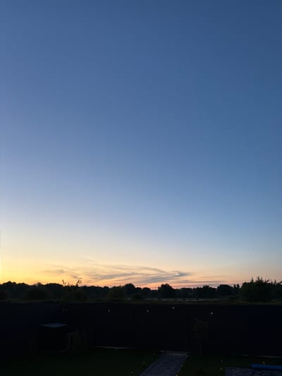 The image shows a twilight sky over a dark, fenced yard. The sky transitions from a deep blue at the top to a soft orange and yellow glow near the horizon, indicating either sunrise or sunset. A silhouetted treeline forms a dark band across the middle ground, with the fence appearing as a stark black barrier in the foreground. The yard is sparsely furnished, with some dark structures and landscaping visible. The location is identified as Sharovechka, Ukraine.