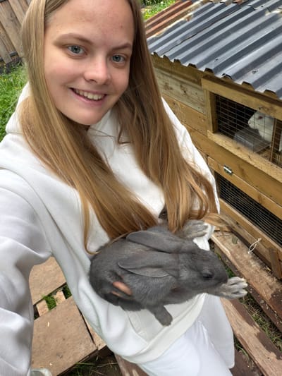 A young woman is depicted outdoors, holding a gray rabbit. She has long, light blonde hair and blue eyes, and is smiling directly at the camera. She is wearing a white sweatshirt. The gray rabbit, characterized by its floppy ears, is comfortably resting in her arms or lap, with one front paw gently raised.

In the background, a rustic wooden hutch or enclosure is visible. Inside one of its compartments, a white rabbit with red eyes can be seen peering out. The structure is partially covered by a corrugated metal roof. The ground features green grass interspersed with wooden planks or pallets.

The photograph captures a bright daytime scene, indicating clear or bright overcast weather. The woman's expression conveys happiness and warmth. The setting suggests a rural or backyard environment, consistent with the location context of Vozhoiskii, Russia. There is no visible text within the image.