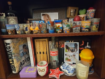 The scene depicts a two-tiered wooden bookshelf filled with personal items, souvenirs, and books, located in the Cherwell District, United Kingdom.
On the top shelf, approximately 15-20 souvenir shot glasses from various international locations (e.g., Dublin, Salzburg, Ibiza 2014, Hawaii, Frankfurt, Rio de Janeiro, Rome) are arranged across the front. Behind these, a dark bottle with a cork stopper, labeled "MONKEY 47," stands next to a small white deer figurine. Further back, a framed photograph shows a woman's face. Other objects include a small framed teddy bear photograph, a green heart-shaped ornament, a red apple-shaped ornament, and a colorful patterned cup.
The bottom shelf contains a large book titled "ADVENTURE" with a world map design, next to a book titled "Cocktail Edit." A framed photograph of a man embracing a woman is placed against the "ADVENTURE" book. A wooden box with three cutouts is visible. A classic glass Coca-Cola bottle stands in front of several dark-spined books, one of which displays a white chess piece (queen or king) on its spine. A vertical photograph of a Siamese cat is positioned prominently. To the right, a yellow ice cream cone-shaped object, a purple rectangular tin with a "Friends" logo, a Santa Claus-themed decorative holder, a red star-shaped ornament inscribed "KEEP CALM and Merry Christmas," two miniature blue and white Dutch clogs, and a glass candle jar with white wax (label partly visible as "MUMFORD") complete the display.
The items are static, arranged for display on the dark wooden shelves. No living individuals are present, only photographic representations.