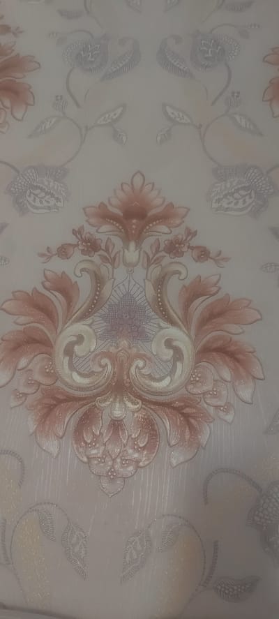 The image displays a close-up view of ornate wallpaper with a repeating damask pattern. The dominant colors are muted peach, cream, and light gray, with subtle shimmering accents. The pattern features elaborate floral motifs and swirling scrollwork, creating a sense of depth and texture. The wallpaper appears to be a decorative element within an interior space, likely a room in a residential or commercial building. There are no people, actions, or specific environmental details beyond the wallpaper itself visible in the image. The location in Ынтымак, Kyrgyzstan, is not discernible from the visual content of the photograph.