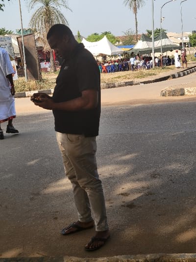 The scene is situated in the city of San Martín, Colombia. A dark-skinned adult male, facing left in profile, stands on a paved road surface with a dirt shoulder. He wears a black polo shirt, light-colored slim-fit trousers, and black flip-flops. He is actively engaged with a small, dark-colored object, likely a smartphone, held in both hands at waist level, with his head tilted downwards. To his immediate left, partially visible, is another individual in a white, loose-fitting garment and dark shoes, moving away from the viewer.
In the background, across a concrete curb and a grassy median, an outdoor gathering is taking place. Numerous individuals are seated on blue plastic chairs under two large tents, one white and one striped green and white. Other people are standing around the periphery of the gathering. Several tall palm trees are visible, along with utility poles and streetlights. Distant buildings indicate an urban or semi-urban setting. The sky is bright, suggesting clear daylight conditions, with shadows cast on the ground indicating strong sun.
