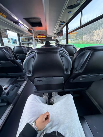 The image depicts the interior of a public transit bus, viewed from a first-person perspective by a seated passenger. The passenger's legs are extended, clad in light grey sweatpants, with feet wearing grey and white patterned athletic shoes visible. A left hand with a silver and gold wristwatch rests on the lap, adjacent to a black fanny pack or crossbody bag.
The bus interior features rows of black upholstered seats with integrated grab handles and fold-down tray tables on their backs. The floor is dark, with silver-colored rails for seat mounting visible along the aisle. The light-colored ceiling contains overhead compartments, air vents, and individual blue-illuminated reading lights or call buttons. A central digital display above the driver's area shows "C 18" in red and Hebrew text: "מגלה עיר חיפה" (Haifa City Explorer). Other informational signs in Hebrew are visible on the bus's left interior wall.
Ahead of the photographer, the back of another passenger's head is visible in the seat directly in front, and further forward, the back of the bus driver's head is discernible. Through the bus's right-side windows, a hilly, vegetated landscape with a light-colored structure behind a bright green fence is visible under an overcast sky.
The scene is located within the city of Nesher, Israel, operating as part of the Haifa metropolitan area transit system.
