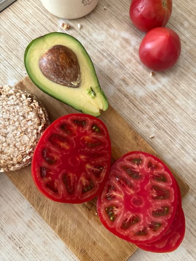 This overhead shot captures a variety of fresh ingredients arranged on a light-colored, textured surface. In the upper left, two whole, ripe red tomatoes are visible. To their right, a wooden cutting board displays sliced red tomatoes, revealing their juicy interior with seeds and pulp. Adjacent to the tomatoes, a halved avocado is positioned, its creamy green flesh contrasting with the rich brown of its pit. At the bottom center, a circular rice cake, textured with grains, sits on the surface. The overall scene suggests the preparation of a meal, likely a sandwich or toast, given the presence of bread-like item, avocado, and tomatoes. The lighting indicates it is daytime, and the setting appears to be a kitchen or dining area. No text is visible in the image.