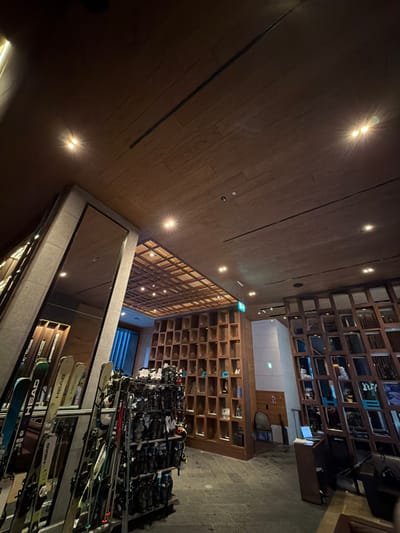 A dimly lit interior view shows rows of shelves filled with shoes and skis. Overhead, a grid-like structure is visible with ceiling lights sporadically illuminating the wooden paneling above. The scene gives the impression of a ski resort or rental facility, with a focus on organization and equipment storage. The perspective is skewed.