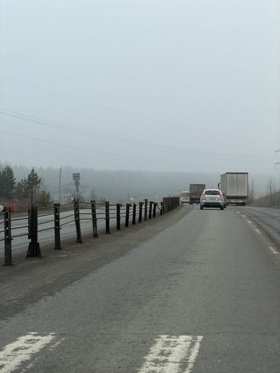 The image depicts a wet, asphalt road on a foggy, overcast day, likely in Bolgury, Russia. The scene suggests poor visibility and damp conditions.

In the foreground, the road surface is dark and appears wet, with visible white lane markings and a solid white line bordering the shoulder on the right. A cable-style guardrail with dark posts and multiple tensioned cables runs along the right side of the road, disappearing into the haze. Some red reflectors are attached to the guardrail posts.

Further down the road, traffic is visible, suggesting slow movement or a queue. A white sedan is prominent among the vehicles, its rear visible, possibly with brake lights illuminated. Its license plate clearly shows "P228BH" followed by two digits, likely indicating a Russian registration. Behind the sedan, several large semi-trailer trucks are seen, their white or light-colored trailers standing out against the muted background. Other vehicles are faintly visible deeper in the fog.

To the left of the road, the landscape is shrouded in dense fog, with bare or sparsely-leaved trees lining the roadside, hinting at late autumn, winter, or early spring. Several utility poles with power lines stretch across the frame on the left. A couple of road signs are visible through the fog on the left side of the road; one appears to be a yellow diamond-shaped warning sign (potentially for an intersection or crossing), and another circular sign is also present. The sky is a uniform, light gray, blending seamlessly with the fog and distant landscape, creating a subdued and monochrome atmosphere. The overall impression is one of a cold, damp, and misty day on a rural highway.