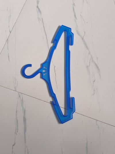 A blue plastic clothes hanger is placed on a floor with white tiles that have faint gray veining. The hanger is oriented diagonally from the bottom left to the top right of the frame. It is a standard adult-sized hanger with a hook at the top and two notches on the sides. The surface it rests on appears to be indoors, with no visible outdoor elements. The lighting is even, and the image is sharp. The specific location in Cavite City, Philippines, cannot be determined from the image.