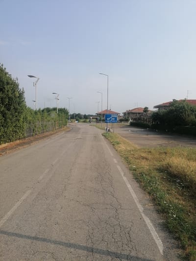 This is a street-level, eye-level, medium shot photograph taken on a clear day in Oleggio, Italy. The camera is positioned in the middle of an asphalt road that recedes into the distance. The road is cracked and worn in places, with a white line marking the edge on the right side. On the left side of the road, there is a dense, tall green hedge behind a metal fence, followed by a line of modern streetlights with solar panels. Several more streetlights are visible further down the road. To the right of the road, there is a patch of dry, tall grass with some small white flowers, and further back, several residential buildings with red-tiled roofs and some trees. A blue directional sign is visible on the right side of the road, indicating directions to "GALLARATE," "OLEGGIO," and "BUSTO A." Several traffic signs are also present near the directional sign. The sky is a pale blue with no clouds. The overall scene depicts a somewhat rural or suburban setting with an aging road and modern infrastructure like streetlights.