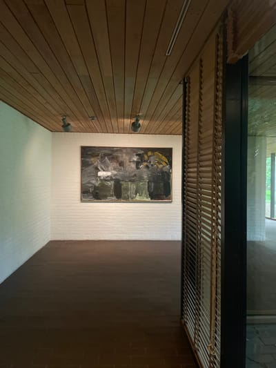 The image displays an interior view of a gallery space, consistent with the architecture of the Louisiana Museum of Modern Art in Humlebæk, Denmark. The room features white painted brick walls and a light brown wooden slatted ceiling. Two gray metallic track spotlights are mounted on the ceiling, directed towards a large, rectangular abstract painting centered on the far wall. The artwork, framed in dark material, exhibits a palette of dark gray, black, olive green, white, and yellow. The floor is covered with dark brown rectangular tiles, likely brick or terracotta, laid in a stretcher bond pattern. To the right, a tall window with horizontal wooden blinds (louvers) is partially visible; the blinds are pulled up and secured. Through the window, an adjacent interior space with white brick walls and similar flooring is reflected, with green foliage from an outdoor environment discernible beyond. No individuals, specific actions, or additional subjects are present in the scene.
