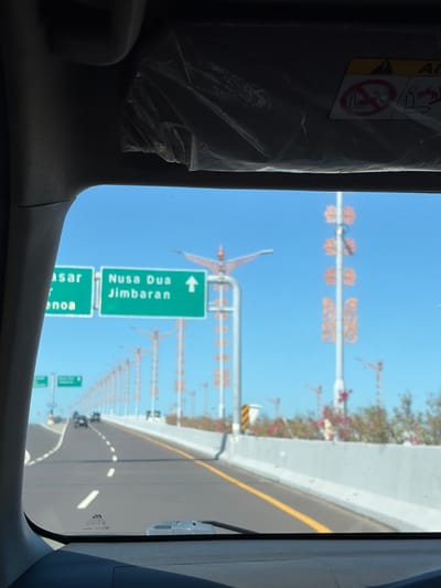 This image captures a view from inside a vehicle traveling on a highway in South Denpasar, Indonesia. The road ahead curves slightly to the left, with dashed white lines marking the lanes and a solid yellow line on the right edge. In the foreground, the dashboard and windshield wipers are visible.
Two green highway signs are prominently displayed on the left side of the road. The left sign appears to indicate destinations such as "Pasar" and "enọa". The right sign clearly reads "Nusa Dua Jimbaran" with an upward-pointing arrow, suggesting these locations are straight ahead.
In the background, a clear blue sky dominates the scene. Numerous decorative lampposts with orange floral motifs line the highway. Some of these lampposts are tall and slender, while others are shorter and sturdier. In the distance, several construction cranes are visible, indicating ongoing development or infrastructure projects. A few vehicles, including cars and possibly a motorcycle, are on the highway ahead. Vegetation with pink flowers is present along the right side of the road, beyond a concrete barrier. The overall environment suggests a sunny day, likely during daylight hours.