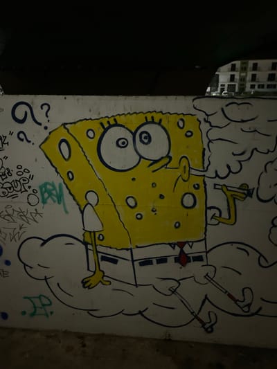 The image shows a graffiti artwork on a wall in Tavira, Portugal. The graffiti depicts SpongeBob SquarePants smoking from two cigarettes while seated on a cloud. He is wearing his signature square pants, white shirt, and red tie. The artwork is done in a cartoonish style with bold outlines and yellow coloring for SpongeBob. In the background, there is other graffiti in various colors and styles, as well as a glimpse of apartment buildings. The scene appears to be an outdoor urban setting, possibly under a bridge or overpass given the dark ceiling visible at the top of the image.