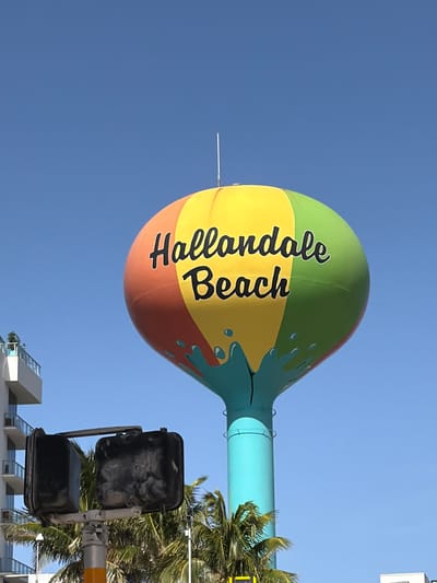 A vibrant, colorful water tower with the words "Hallandale Beach" painted in black, cursive script stands tall against a clear blue sky in Hallandale Beach, Florida, United States of America. The water tower is painted with broad stripes of red, yellow, and green, and features a teal base with a design resembling splashing water. To the left of the water tower, a modern building with balconies and a white facade is partially visible. In the foreground, a traffic signal with black casings is mounted on a pole. Palm trees with lush green fronds are visible behind the traffic signal and around the base of the water tower, indicating a tropical or subtropical environment. The overall scene suggests a sunny day in a coastal city.