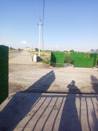 The image depicts an outdoor scene with a paved area in the foreground and a road receding into the distance. Artificial green fences are placed on the right side of the image, some with a shallow pool of water in front of them and chairs behind. A shadow of a person holding a phone is cast on the pavement in the foreground, along with the shadow of a fence. The environment includes a clear blue sky, power lines, and vegetation. The road shows some traffic and buildings in the background. The overall impression is of a somewhat rustic or underdeveloped area with elements of artificial landscaping. The location is specified as Shar-Bakr, Uzbekistan.