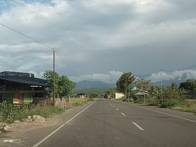 The image captures a roadside scene in Keude Ulim, Indonesia, characterized by a paved asphalt road with white dashed lines indicating lanes. The sky is overcast with heavy, dark clouds, suggesting impending rain. On the left side of the road, a simple structure with a dark, possibly corrugated, roof and wooden fencing is visible, bordered by lush green vegetation and scattered rocks. Further down the road, a yellow construction vehicle, possibly a bulldozer or loader, is stationary in the distance. On the right side of the road, there are trees, smaller buildings, and dense greenery. The background features rolling hills and mountains partially obscured by the cloudy sky. The overall impression is one of a rural or semi-rural environment with ongoing construction or maintenance activities. The watermark "anroja" is present in the bottom left corner of the image.
