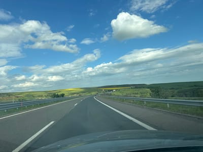 A low-angle perspective shows a straight stretch of asphalt highway with two lanes in each direction, divided by a solid white line and featuring white reflector posts along the edges. The highway curves slightly to the right in the distance. A metal guardrail runs along both sides of the road, separating it from grassy terrain with rolling hills. The sky is blue with scattered cumulus clouds. In the distance, there is a field of yellow flowers. The bottom of the image shows the hood of a car, suggesting the photo was taken from inside a vehicle. The location appears to be near Vratitsa, Bulgaria, based on the landscape and road infrastructure. No people are visible in the image. The subjects are the highway and surrounding landscape. The objects include cars on the horizon, guardrails, and vegetation. The action is the movement of the vehicle on the highway. The environment is rural with a mix of cultivated fields and natural vegetation.
