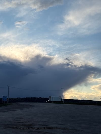 The image presents a view in Chernovskoe, Russia. The sky dominates the scene, displaying a stark contrast between a patch of blue sky with scattered white clouds and an approaching dark, ominous storm cloud. A section of pavement is visible in the foreground. In the middle ground, there's a narrow strip of dense, dark green forest. An electric pole and some road signs stand near a white van. The lighting is uneven, with the part of the sky unaffected by the storm illuminated, casting shadows onto the landscape.