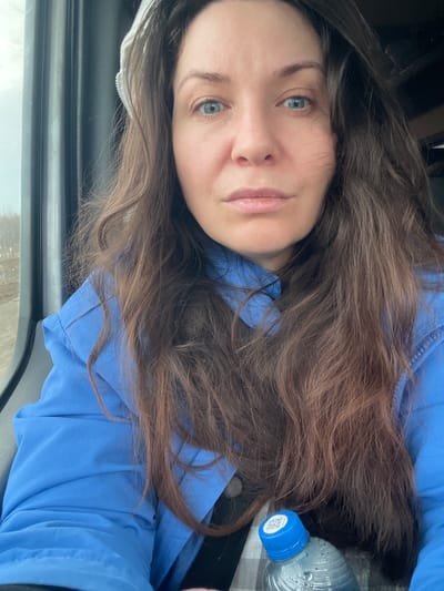 A woman with long brown hair and blue eyes is pictured indoors, wearing a blue shirt. A water bottle with a blue cap is visible to her right. The scene is possibly inside a vehicle due to the presence of a car window frame. There is no information in the image to determine location in Yus'ki, Russia.