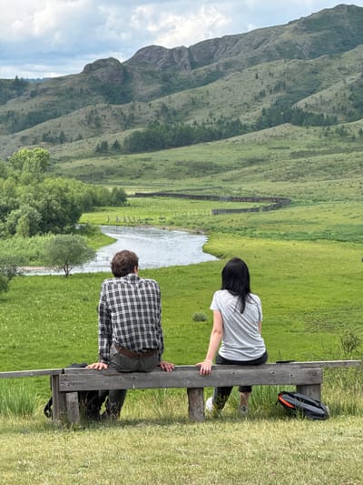 Two individuals are seated on a wooden bench overlooking a verdant valley in Anchyl-Chon, Russia. The person on the left, with brown hair, is wearing a plaid long-sleeved shirt and dark pants. The person on the right, with black hair, is wearing a grey t-shirt and dark pants. A black backpack is positioned on the ground to the left of the bench, and another bag, primarily orange and black, is on the ground to the right.
The landscape features rolling green hills and mountains in the background, with a winding river flowing through the valley in the middle ground. A wooden fence runs along the riverbank. The sky is overcast with scattered clouds. The bench is situated on a grassy incline in the foreground.