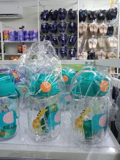 The scene depicts the interior of a retail establishment, presumed to be in the city of Kauman, Indonesia. In the immediate foreground, multiple clear plastic water bottles are displayed on a white counter. Each bottle is individually encased in a clear plastic bag, features a teal green lid with an orange push-button mechanism, and is decorated with cartoon depictions of a light blue elephant and a yellow giraffe. A green straw is visible inside several of these bottles.
In the background, two white metal shelving units are visible. The left unit displays various merchandise: the top shelf contains rows of blue cylindrical objects, while the middle shelf holds orange and black cylindrical containers. Partially visible green potted plants are on a lower shelf. Above this unit, a white wall-mounted air conditioning unit is installed. The right shelving unit displays numerous pairs of sandals or flip-flops, sorted by color and suspended on white plastic hangers. The left portion of this unit exhibits dark blue sandals, while the right portion features black, beige, and lighter-toned sandals. No individuals are present, and no actions or interactions are depicted.