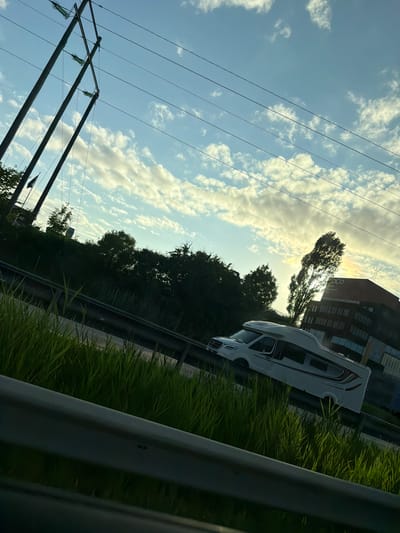 The image depicts a white camper van driving on a highway in Mölndal, Sweden, likely during late afternoon or early evening. Tall power lines are visible in the sky, with multiple wires suspended between two large wooden poles. The sky is a light blue, with scattered white and yellowish clouds, suggesting the sun is low on the horizon.
In the foreground, lush green grass grows alongside the highway, separated by a metal guardrail. Beyond the highway, a dense line of trees forms a dark green barrier. To the right of the frame, a modern brick building with large windows is visible, bearing the text "COCO" on its facade, indicating it's part of a business or innovation park. A tall, slender tree stands in front of the building, silhouetted against the brighter sky. The camper van is the primary subject in motion, traveling on the highway. No people are clearly visible within or around the van. The overall scene suggests a journey through an urban or suburban area with interspersed natural elements.